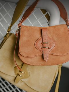 Due borse in pelle scamosciata, una marrone chiaro e una beige, con cinturini in pelle e chiusure dorate, appese su uno sfondo di griglia metallica bianca.