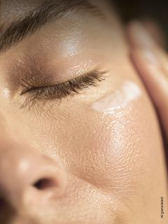 Un primo piano del viso di una persona con gli occhi chiusi, che mostra una pelle liscia e luminosa e un tocco di crema sotto l'occhio.