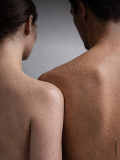 Una donna e un uomo sono in piedi schiena contro schiena, entrambi senza camicia, e mostrano le loro spalle e schiene lentigginose su uno sfondo neutro.