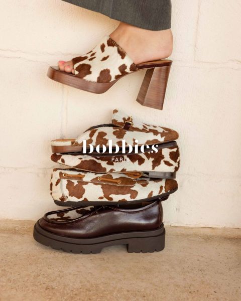 Quattro scarpe alla moda, tra cui tacchi con stampa mucca, mocassini e una scarpa grossa marrone scuro, impilate davanti a un muro chiaro con sovrapposizione di testo "Bobbies Paris".