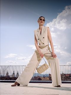 Una donna in un tailleur pantalone beige senza maniche e con occhiali da sole posa all'aperto, con una borsa abbinata in mano, con un paesaggio urbano e un cielo blu sullo sfondo.