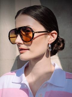 Una donna con capelli castani raccolti in uno chignon basso indossa grandi occhiali da sole color ambra, orecchini d'argento e una camicia rosa e bianca con colletto, illuminata dalla luce del sole.