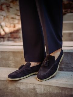 Persona con scarpe scamosciate scure con nappine e pantaloni scuri, in piedi su una scala di cemento all'aperto con uno sfondo sfocato di muro di mattoni.