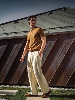Un uomo con una polo marrone e pantaloni color crema a gamba larga esce vicino a un edificio moderno con un tetto metallico inclinato.