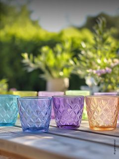 Quattro bicchieri di vetro colorati e strutturati sono su un tavolo di legno all'aperto, con piante verdi sfocate e luce solare sullo sfondo.