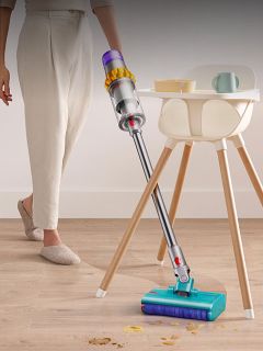 Una persona usa un aspirapolvere senza fili per pulire un pavimento in legno dalle briciole vicino a un seggiolone e un tavolino con delle stoviglie.