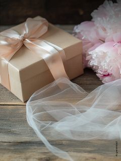 Scatola regalo con nastro rosa pallido accanto a peonie rosa pallido e tessuto bianco su una superficie di legno.