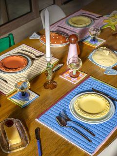 Un tavolo da pranzo in legno con piatti colorati, tovagliette trapuntate, bicchieri, posate e candele in una composizione luminosa ed eclettica.