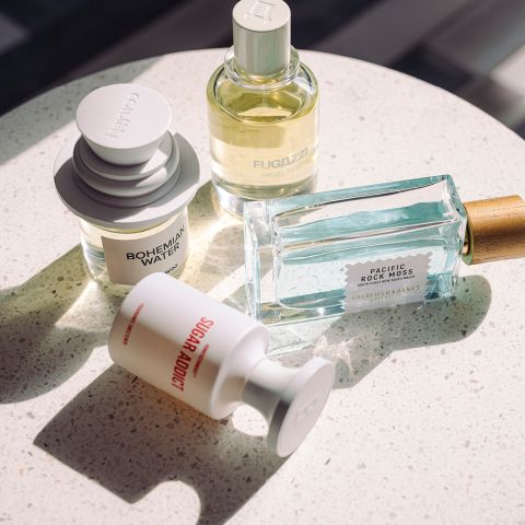 Quattro diverse bottiglie di profumo su una superficie bianca screziata, illuminate dalla luce naturale del sole, ognuna con forme ed etichette uniche.