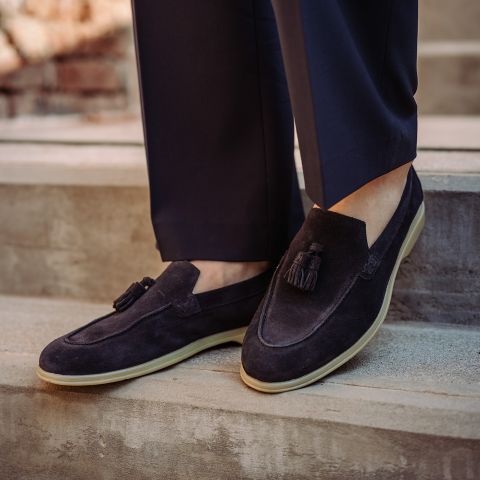 Persona con mocassini con nappine in pelle scamosciata blu scuro con suole color crema, abbinati a pantaloni blu navy, in piedi su gradini di pietra.