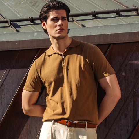 Un uomo con una polo marrone e pantaloni beige è in piedi all'aperto alla luce del sole, con una mano in tasca, di fronte a una struttura metallica.