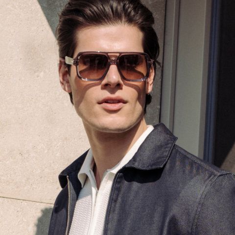 Un uomo con grandi occhiali da sole marroni e una giacca scura è all'aperto alla luce del sole e guarda leggermente di lato.