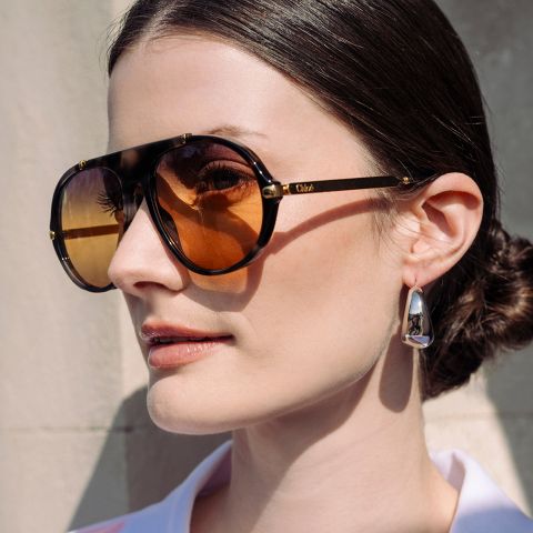 Donna con capelli scuri raccolti in uno chignon basso, che indossa occhiali da sole marroni oversize e orecchini d'argento, in piedi davanti a una parete chiara.