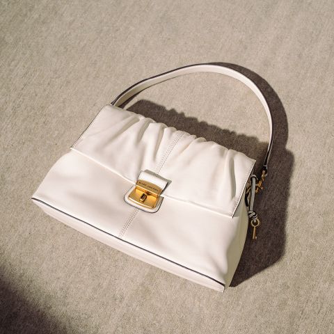 Borsa color crema con fibbia dorata e manico semplice, appoggiata su una superficie di cemento chiaro alla luce naturale del sole.