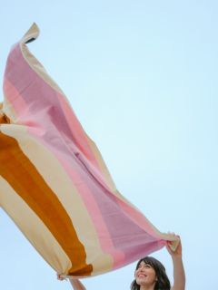 Una donna sorride e tiene alta una grande coperta a righe colorate davanti a un cielo azzurro limpido.