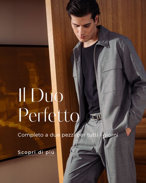 Un uomo in un elegante abito grigio a due pezzi e una camicia nera è in piedi in una casa vicino a una porta di legno e guarda in basso. Il testo recita: "Il Duo Perfetto.