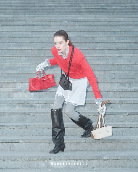 Una donna con un top rosso, guanti grigi e stivali neri scende una scalinata in pietra, con in mano una borsa rossa e una beige, sotto il testo "Margesherwood".