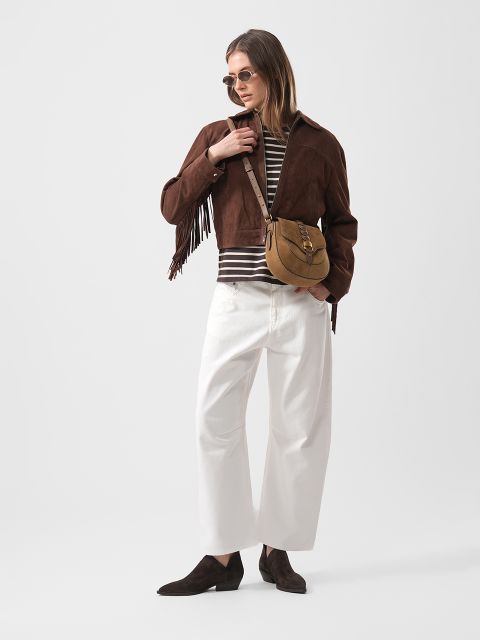 Donna in giacca di pelle scamosciata marrone con frange, camicia a righe, pantaloni bianchi a gamba larga, scarpe marroni, occhiali da sole e borsa a tracolla marrone chiaro, in piedi con sicurezza.