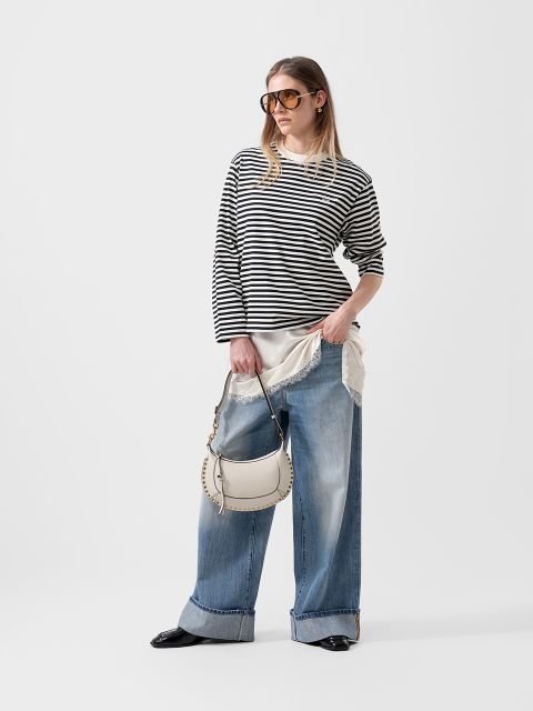 Donna con un maglione a righe bianche e nere, jeans a gamba larga, occhiali da sole e borsa bianca davanti a uno sfondo bianco semplice.