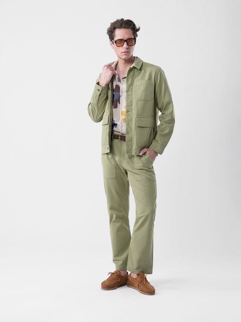 Un uomo che indossa occhiali da sole, una giacca e pantaloni verde chiaro, una camicia fantasia e scarpe marroni, è in piedi davanti a uno sfondo bianco semplice.