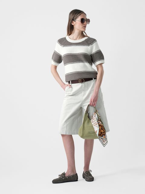 Donna con occhiali da sole che indossa un maglione a maniche corte a righe, pantaloncini bianchi al ginocchio e scarpe basse, e tiene in mano una borsa verde con una sciarpa.