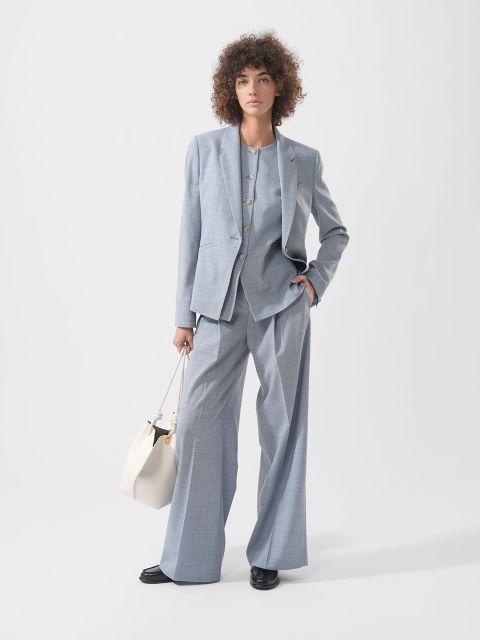 Una donna con i capelli ricci indossa un abito grigio chiaro ampio, scarpe nere e tiene una borsa bianca davanti a uno sfondo bianco semplice.
