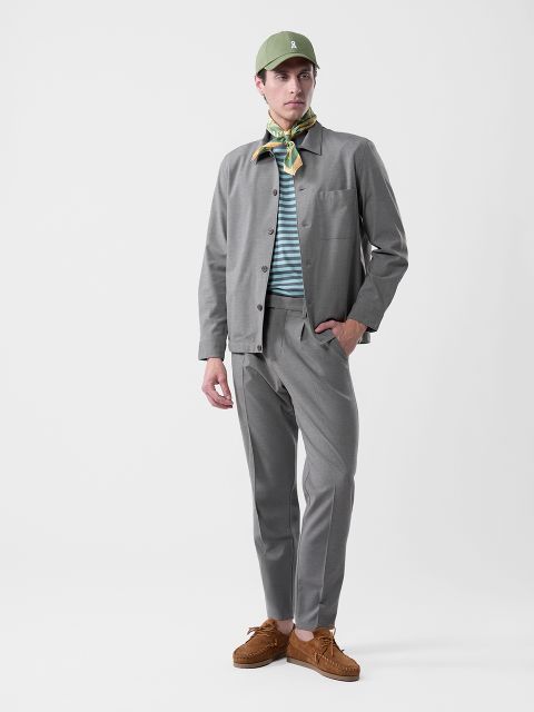 Uomo in abito grigio, camicia a righe, cappello verde, foulard a fantasia e scarpe marroni su uno sfondo bianco semplice.