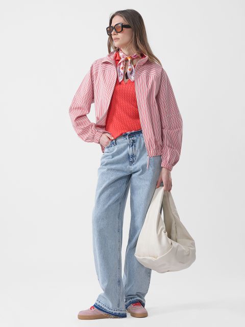 Donna con occhiali da sole indossa una giacca a righe rosa, un maglione rosso, jeans blu e una sciarpa e tiene una grande borsa bianca davanti a uno sfondo bianco semplice.