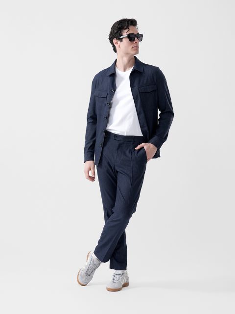 Un uomo che indossa occhiali da sole neri, una giacca blu navy, una maglietta bianca, pantaloni blu navy e scarpe da ginnastica grigie, è in piedi con un piede incrociato davanti all'altro e guarda di lato.