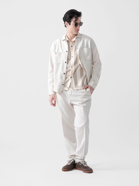 Un uomo con occhiali da sole, giacca beige chiaro, camicia a righe e pantaloni abbinati sta in piedi con le mani in tasca davanti a uno sfondo bianco semplice.