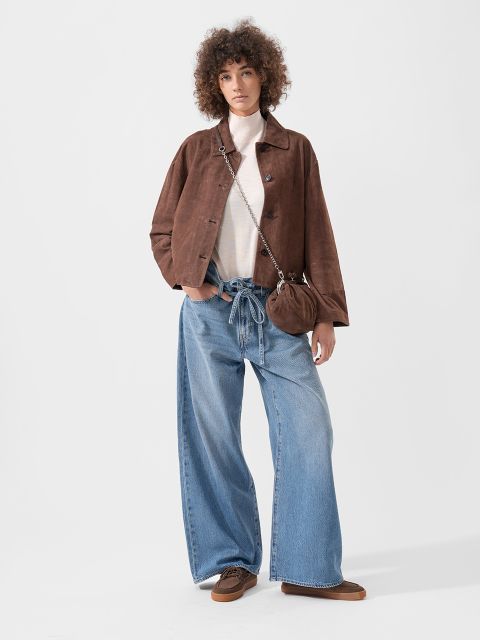 Persona che indossa una giacca marrone, un top bianco, jeans blu larghi, scarpe marroni e una piccola borsetta marrone, in piedi davanti a uno sfondo semplice.