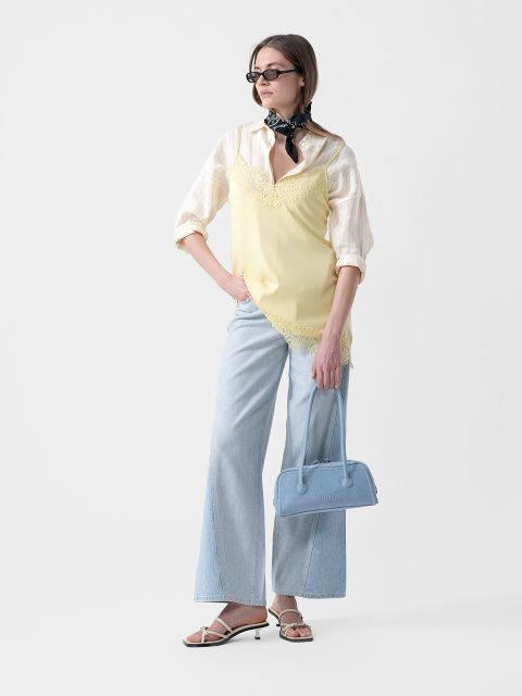Donna con occhiali da sole che indossa una canottiera gialla sopra una camicia a righe, jeans azzurri a gamba larga, sandali e una sciarpa e tiene una borsa blu.