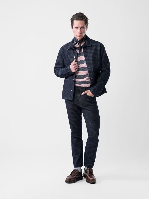 Un uomo posa in una giacca e pantaloni a righe blu navy, una camicia a righe rosa e grigie, scarpe marroni e una sciarpa fantasia davanti a uno sfondo bianco semplice.
