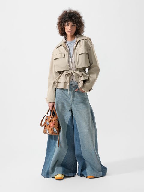 Una persona che indossa una giacca beige, jeans blu larghi e scarpe gialle e tiene in mano una borsa a mano marrone fantasia, è in piedi davanti a uno sfondo bianco.