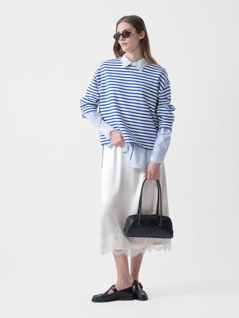 Donna con occhiali da sole che indossa un maglione a righe blu e bianche sopra una camicia con colletto, una gonna bianca con finiture in pizzo, scarpe basse nere e una borsa nera.