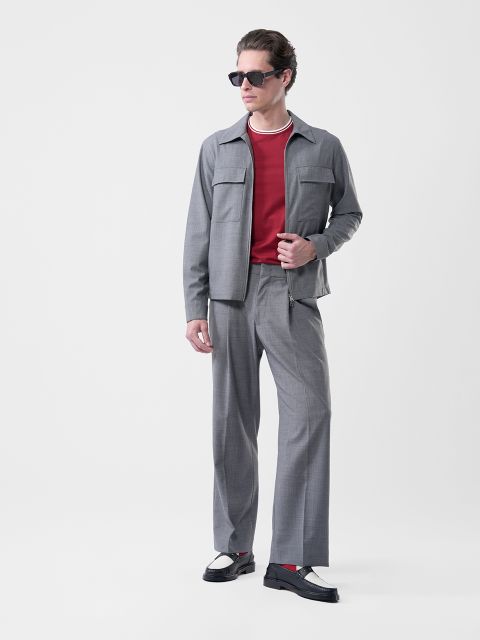 Uomo con occhiali da sole, abito grigio, camicia rossa, scarpe bianche e nere e una collana, su sfondo bianco.