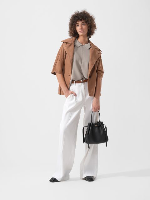 Donna con capelli ricci che indossa una giacca marrone chiaro, una camicia beige e pantaloni bianchi a gamba larga e tiene in mano una borsa nera, in piedi davanti a uno sfondo semplice.