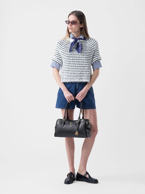 Donna con occhiali da sole che indossa un maglione a righe sopra una camicia con colletto, pantaloncini di jeans e scarpe nere, e tiene una borsa nera davanti a uno sfondo bianco.