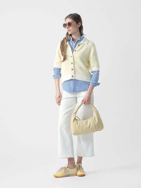 Donna con occhiali da sole, camicia a righe blu, cardigan a righe, pantaloni bianchi e scarpe gialle tiene una borsetta giallo chiaro e si trova di fronte a uno sfondo bianco.