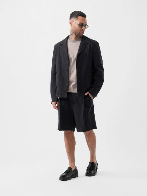 Un uomo con occhiali da sole, giacca nera strutturata, maglietta beige, pantaloncini neri e pantofole nere è in piedi davanti a un semplice sfondo bianco.