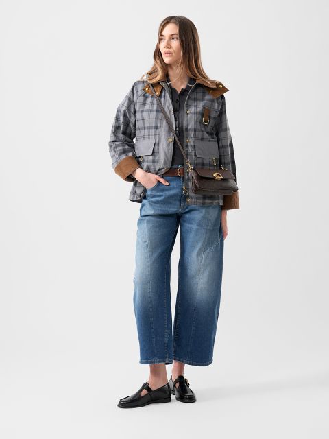 Una donna con una giacca a quadri, jeans blu a gamba larga, scarpe nere e una borsa a tracolla marrone è in piedi davanti a un semplice sfondo bianco.
