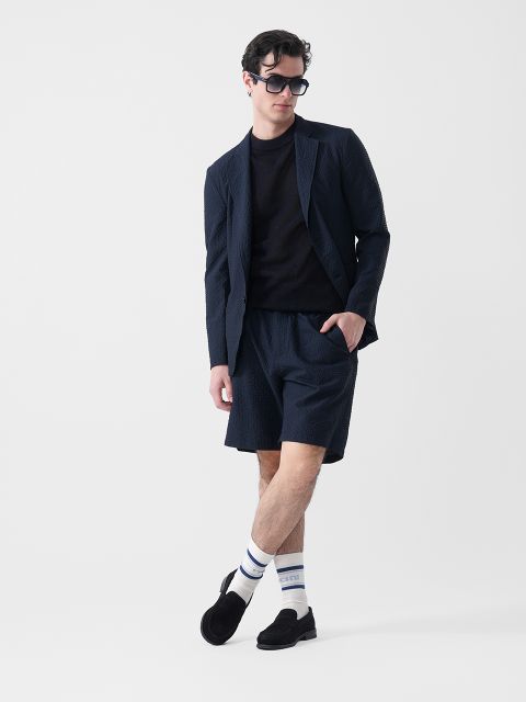 Un uomo che indossa un blazer scuro, pantaloncini abbinati, una t-shirt nera, occhiali da sole, calzini bianchi a righe e pantofole nere, è in piedi davanti a uno sfondo bianco.