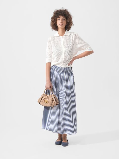 Donna con un top in maglia bianca a maniche corte, una gonna lunga a righe blu e bianche, ballerine blu navy e una borsa beige su uno sfondo semplice.