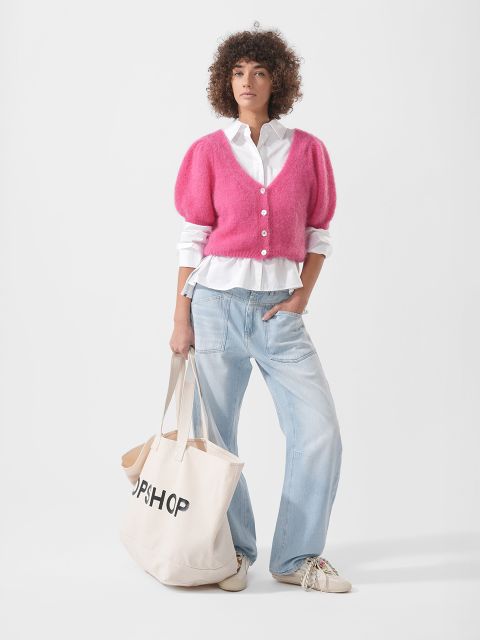 Una persona con cardigan rosa, camicia bianca, jeans azzurri e scarpe da ginnastica tiene in mano una grande borsa tote beige con la scritta "TOPSHOP".