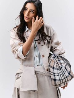 Donna in abiti beige chiaro con una borsa a quadri, che guarda di lato e si tocca i capelli davanti a uno sfondo semplice.