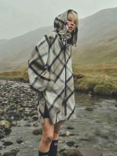 Una persona che indossa un impermeabile a quadri con cappuccio si trova vicino a un ruscello roccioso in un paesaggio nebbioso ed erboso, con colline sullo sfondo.