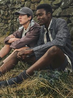 Due uomini siedono in abiti casual da esterno sull'erba secca accanto a un muro di pietra e guardano pensierosi in lontananza.