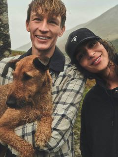 Un uomo sorridente tiene in braccio un cane marrone mentre è in piedi accanto a una donna sorridente con un cappello nero, all'aperto in una giornata nuvolosa.