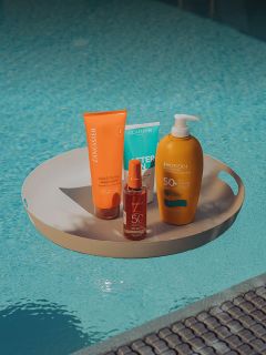 Quattro flaconi di crema solare di diverse marche su un vassoio rotondo che galleggia in una piscina con acqua blu sullo sfondo.