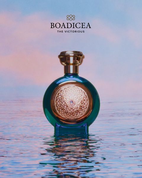 Un flacone di profumo "Boadicea" blu e oro poggia su acqua liscia come uno specchio, sullo sfondo di un cielo color pastello.